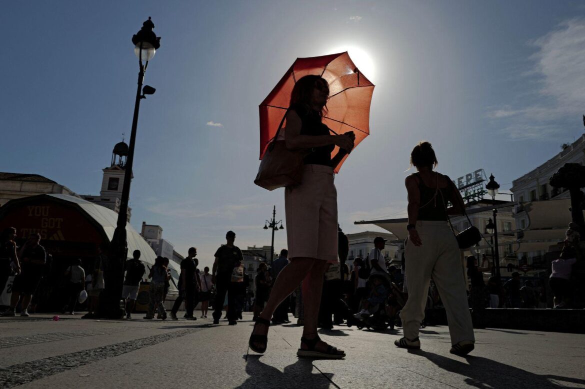 Woman in a black top and pink shorts walks through a busy sunny plaza, carrying a red umbrella that backlights the scene.