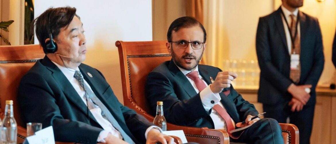 Two suited men seated in a panel discussion; the man on the left wears a headset and listens, while the man on the right speaks and gestures.