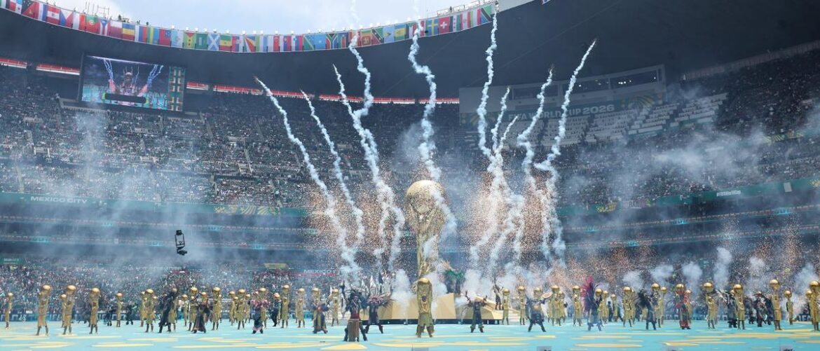 World Cup opening ceremony in a large stadium with a golden trophy centerpiece, performers on a blue field, and fireworks above.