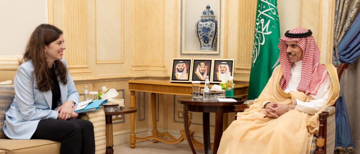 Woman in a light blue blazer speaks with a Saudi official in traditional attire in an ornate room, with portraits and a green flag nearby.