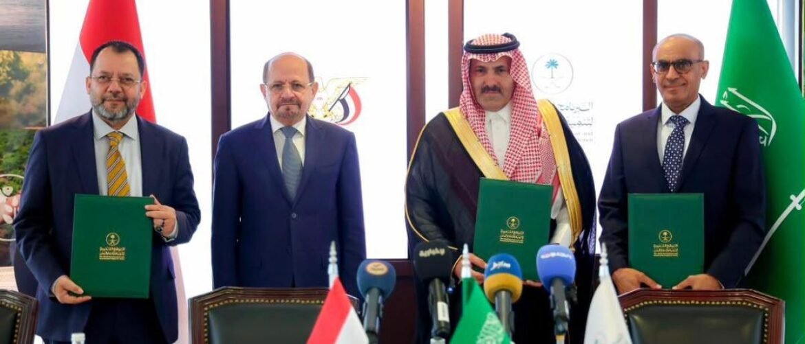 Four men in suits stand behind a table, each holding a green folder as they pose after a signing ceremony with flags and microphones in front of them.