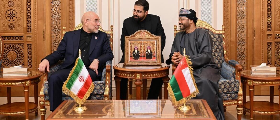 Diplomatic meeting: three men in formal wear sit in an ornate room, with Iranian and Omani flags and a framed icon on a central table.