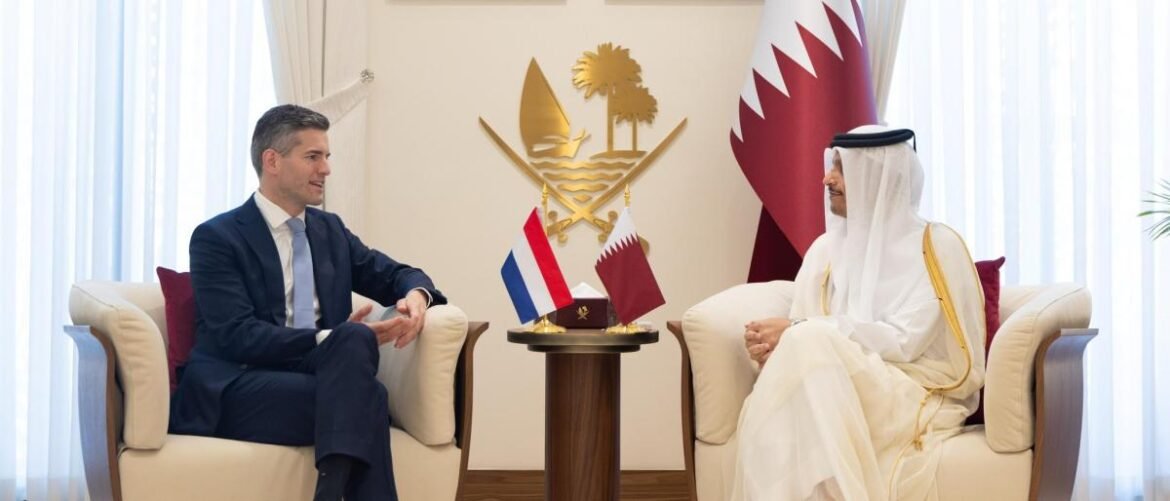 Two men in a formal meeting sit in cream armchairs with a small round table between them; Dutch and Qatari flags are on the table.