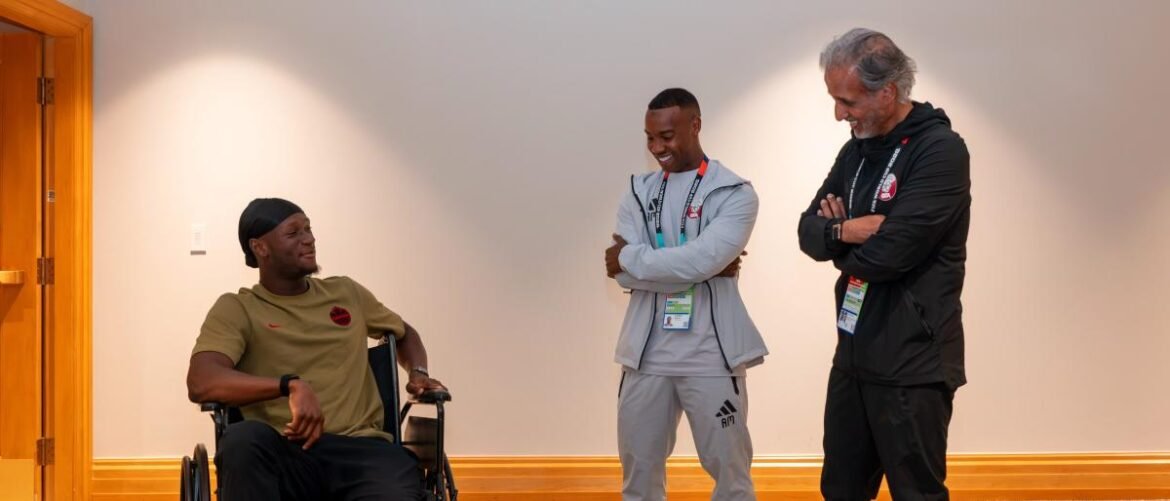 Man in a wheelchair smiles as two men chat and laugh in a bright hallway, creating a friendly moment in a sports setting.