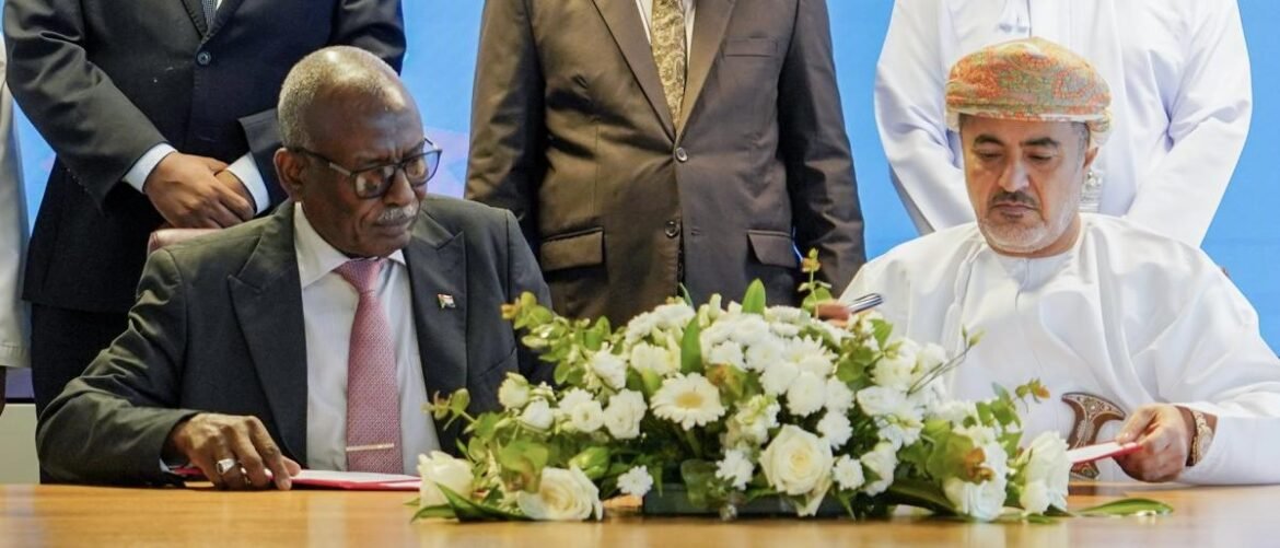 Two men sign documents at a table, one in a dark suit and pink tie, the other in traditional Omani attire, with a white floral centerpiece in front.