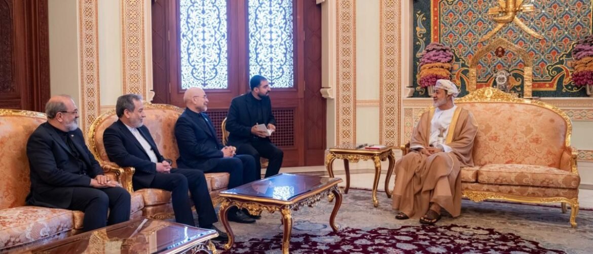 Five men sit on ornate gold-and-cream sofas in a luxurious, ornamented room, chatting with a man in traditional robes and head covering across from them on another sofa.