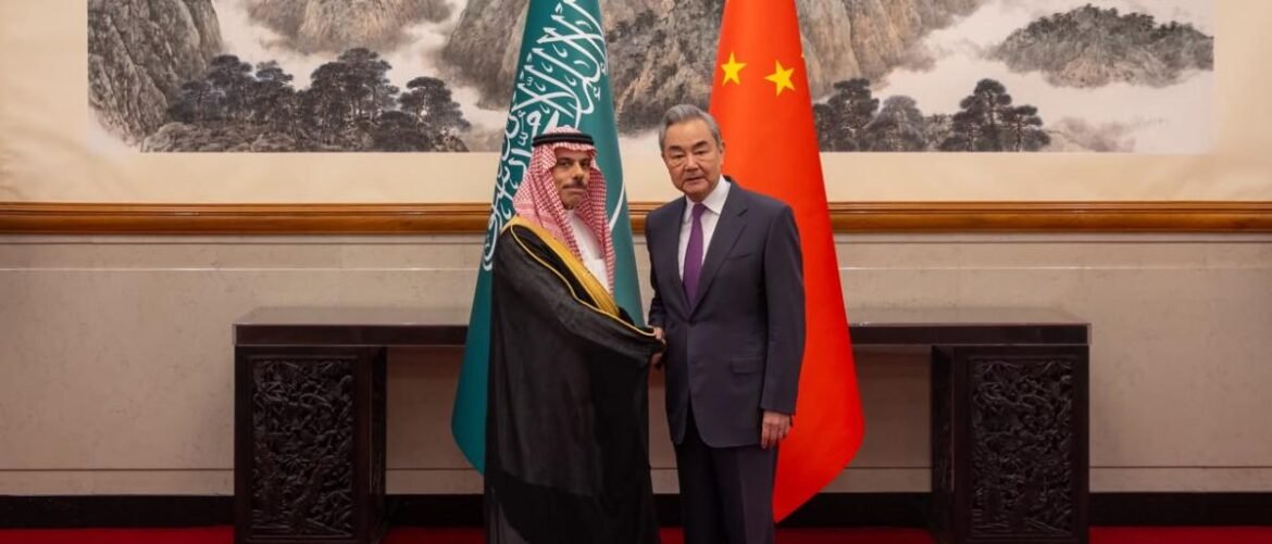 Two male diplomats shake hands in a formal room, flanked by Saudi and Chinese flags in the background.