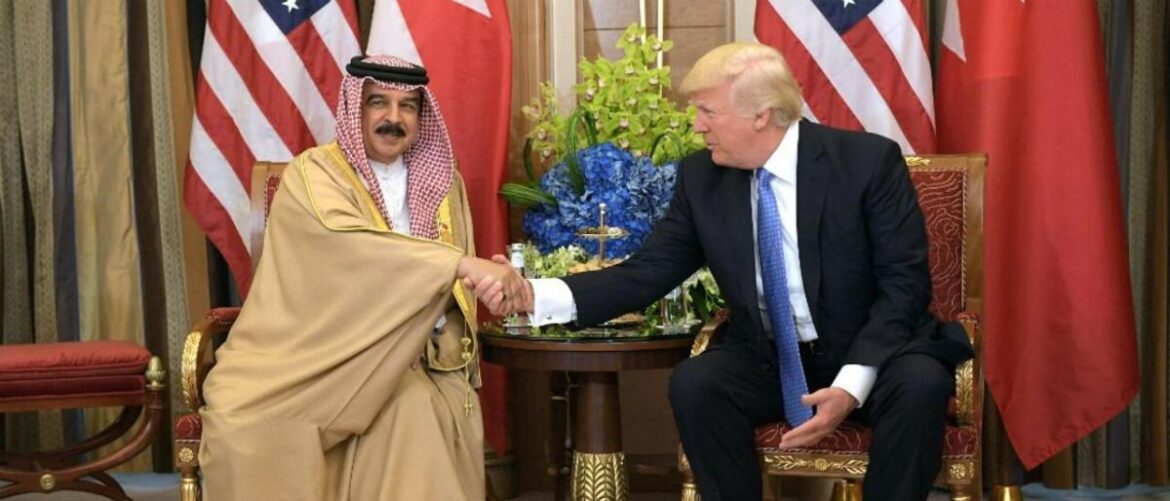Two men in formal dress shake hands in a diplomatic meeting, with US flags and a floral arrangement in the background.