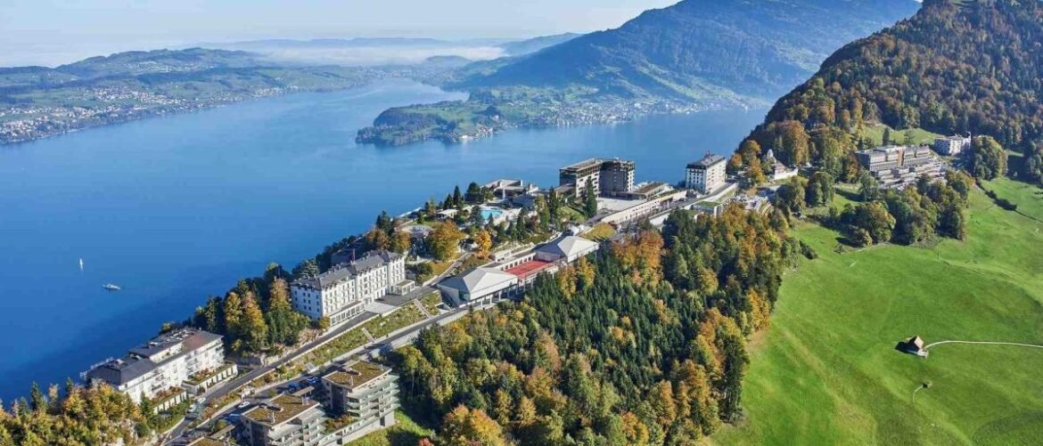 Aerial view of a lakeside town along a hillside with colorful autumn trees, overlooking a calm blue lake and distant mountains.