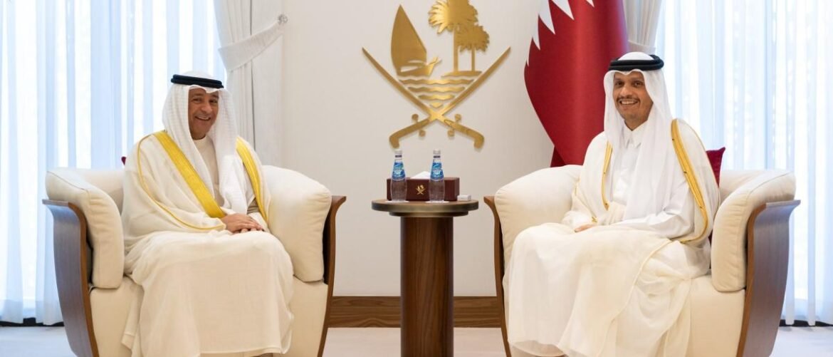 Two men in traditional Qatari attire sit in cream armchairs for a formal meeting, with water bottles on a small table between them and a Qatari emblem behind.