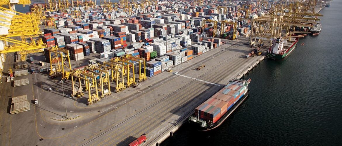 Aerial view of a busy container port with colorful stacked containers, yellow gantry cranes, and ships at the dock along the water.