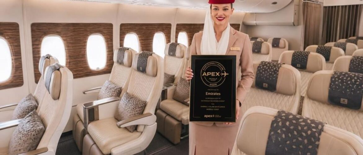 Flight attendant in uniform standing in a cabin, holding an APEX award plaque for Emirates.