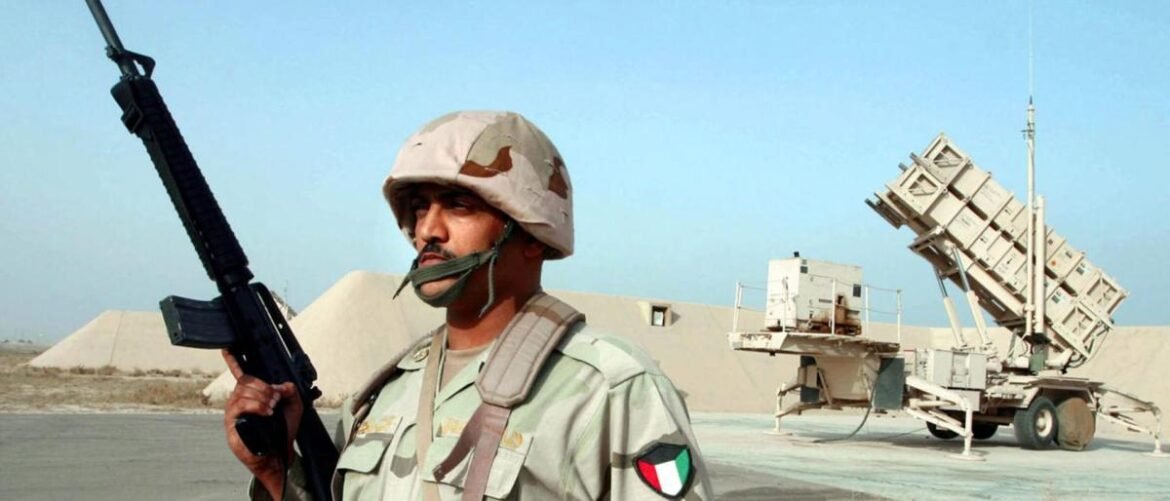 Soldier in desert camouflage holding a rifle with helmet and mask, standing near a large mobile launch system in a sandy base.