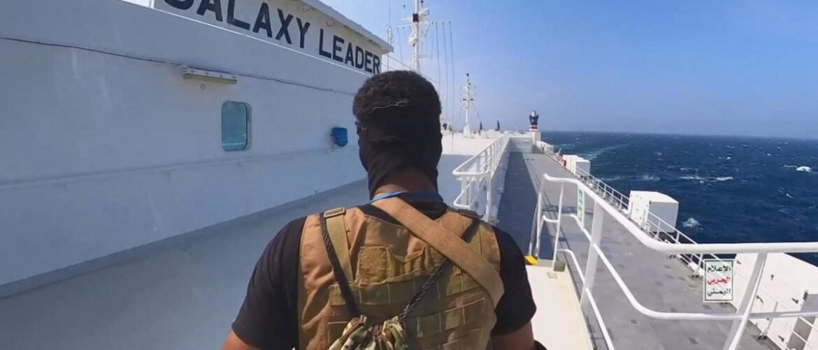 Back view of a person in a brown tactical vest walking on the deck of a white ship with 'GALAXY LEADER' on the hull, ocean to the right.