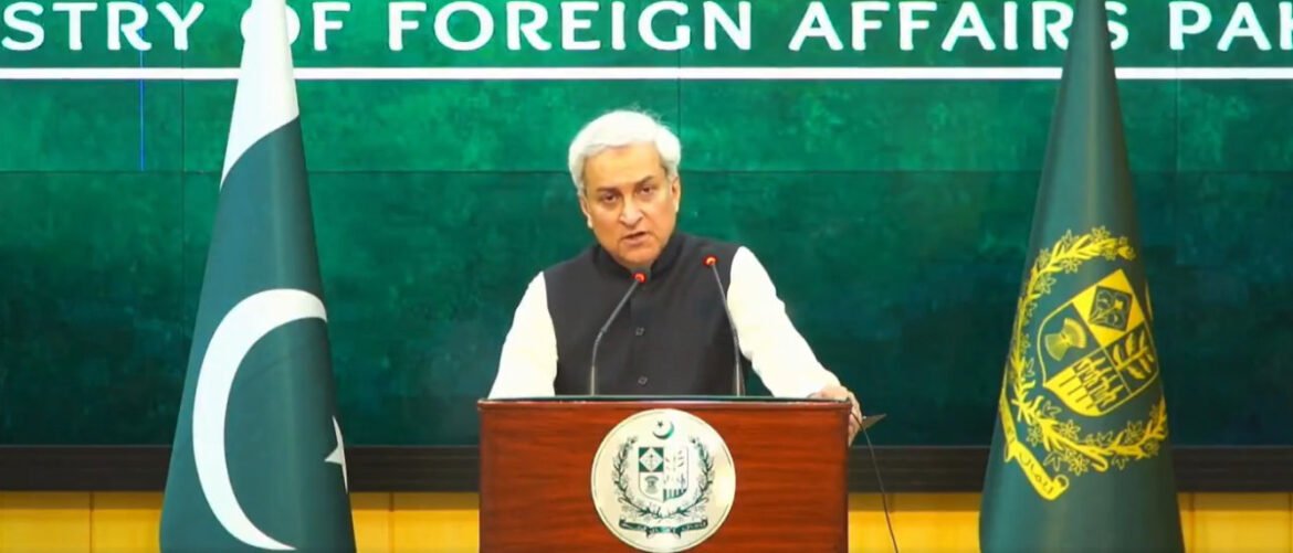 Official press briefing: man speaks at a podium with the Pakistani flag on the left and a government emblem flag on the right, green backdrop reading Ministry of Foreign Affairs.