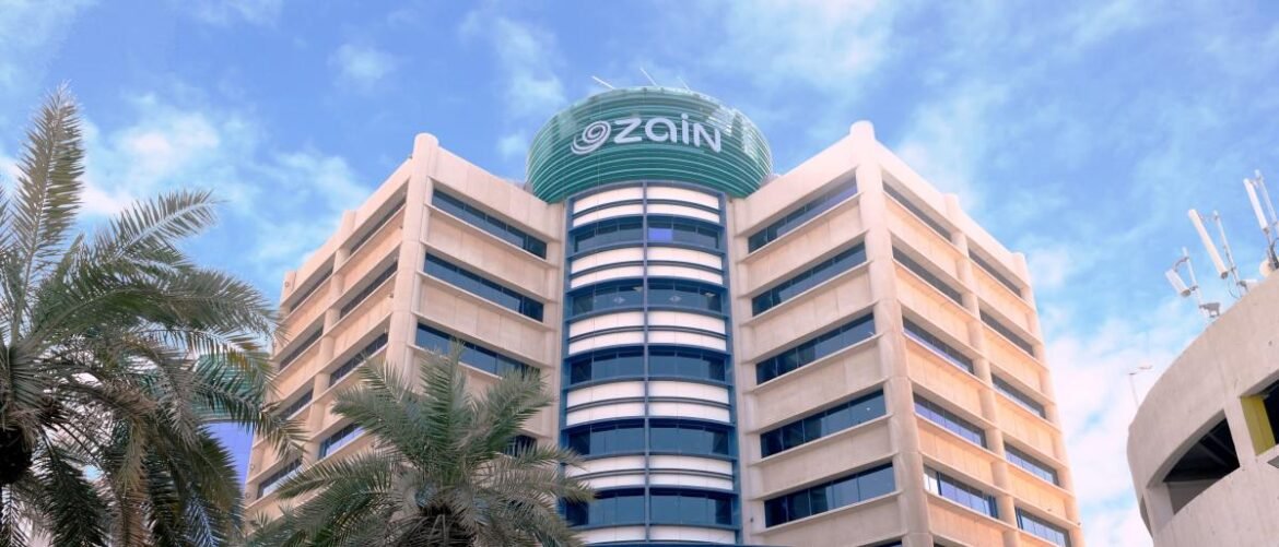 Modern office building with a green circular sign reading 'zain' atop the center tower, blue glass windows, palm trees in foreground.