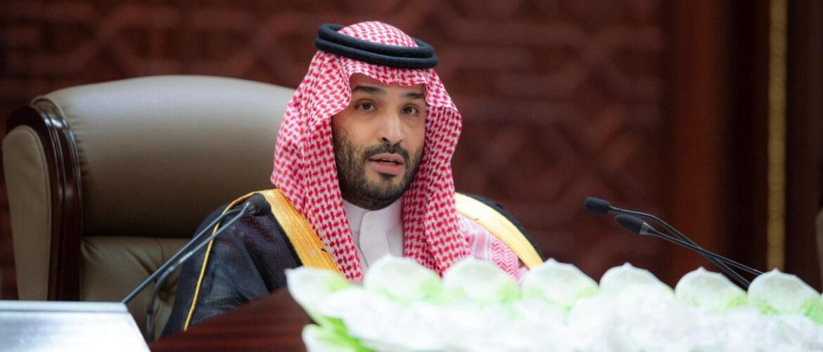 Saudi official in traditional dress sits at a desk with microphones during a formal meeting, facing the audience.