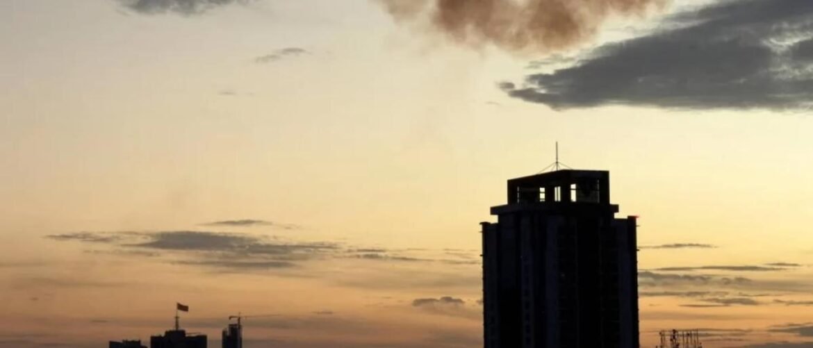 Silhouetted city skyline at sunset with a tall dark building in the foreground and warm orange sky behind it.