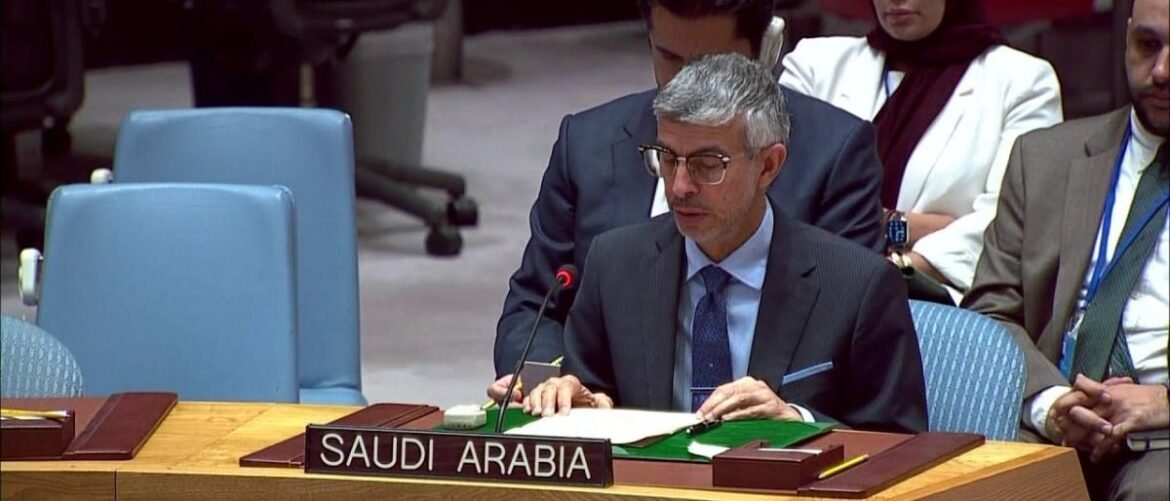Man in a dark suit and glasses signs documents at a Saudi Arabia delegation desk in a formal meeting room.