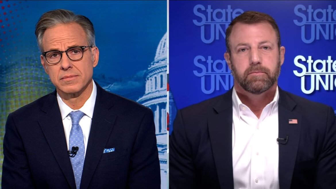 Split-screen news interview showing two men in suits, each in a different studio backdrop, one with glasses and a blue tie on the left, the other with a white shirt and US flag pin on the right.
