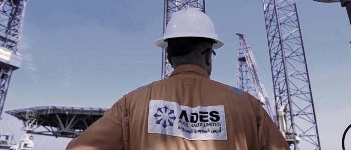 Construction worker in a brown safety jacket and white hard hat, seen from behind, carrying an ADES Saudi Limited patch, with large cranes in a construction site.