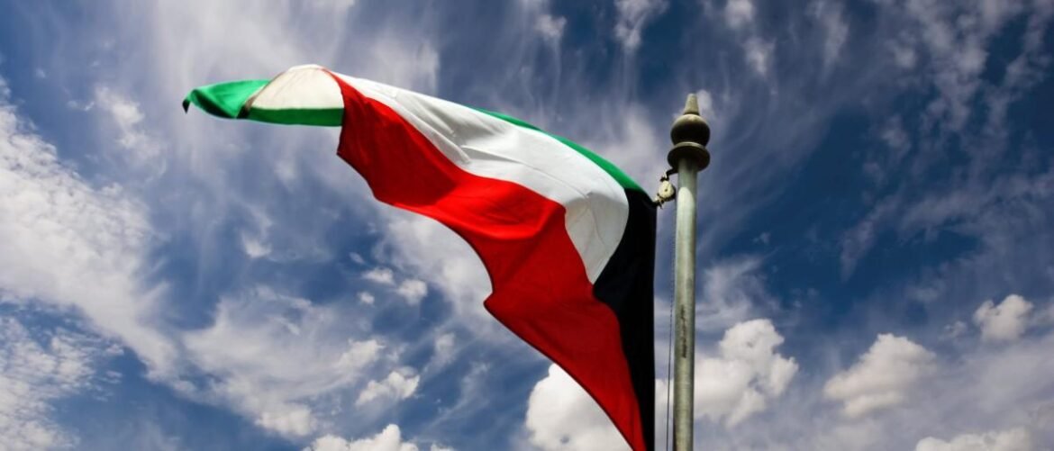 Kuwaiti flag waving on a flagpole against a blue sky with white clouds.