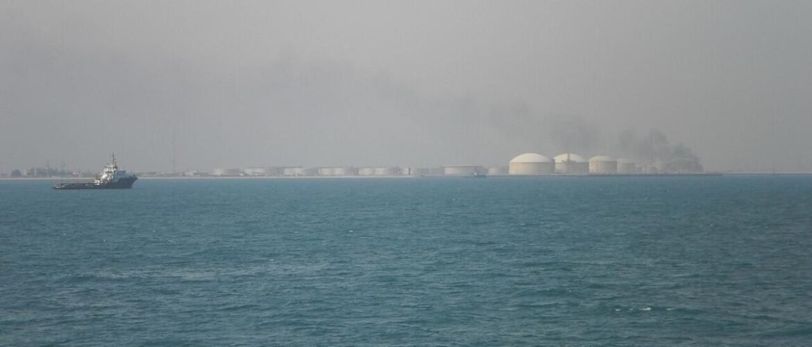 Tugboat on a blue sea near a distant industrial coast with white cylindrical storage tanks and haze-smoked sky.