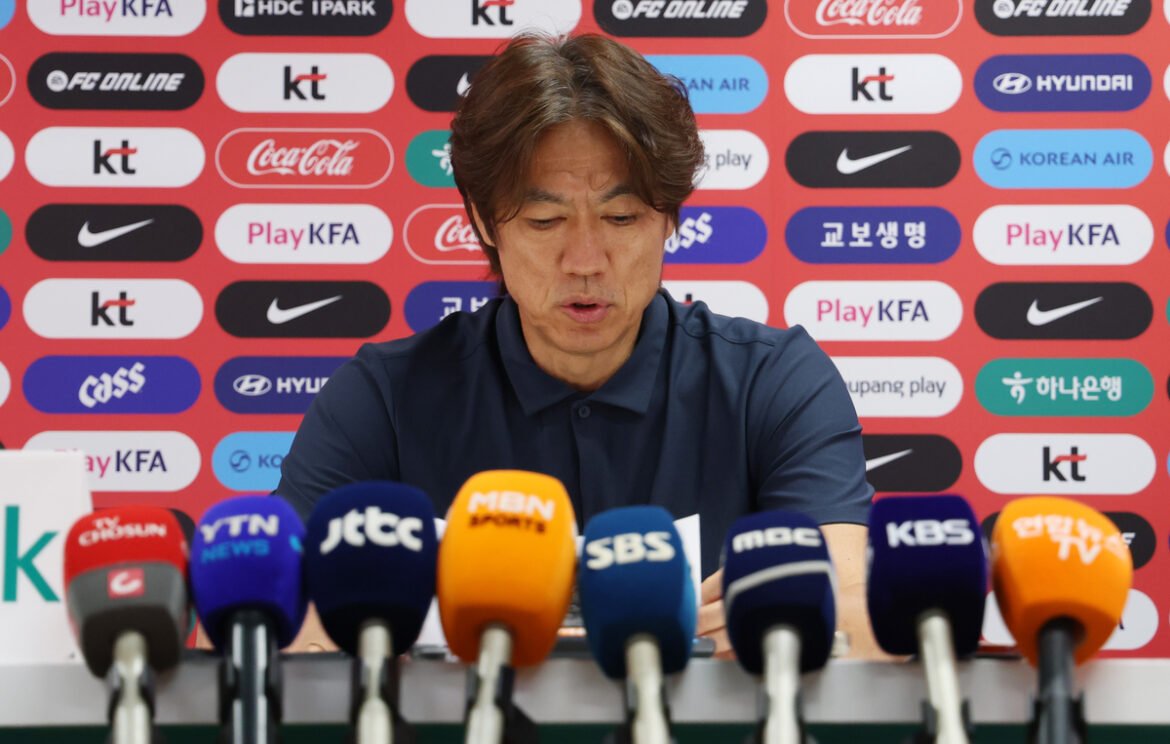 Man in a navy polo speaks at a press conference with multiple branded microphones in front of him on a table.