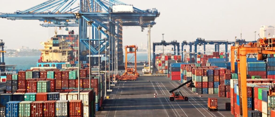 Busy container port with stacked colorful cargo containers and tall blue gantry cranes overhead and along the dock idea of ships nearby