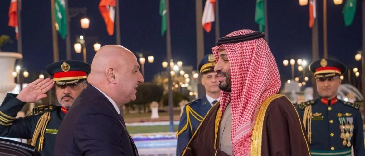 Two men greet each other at night; one in a suit, the other in traditional Saudi attire, with uniformed officers saluting in the background.