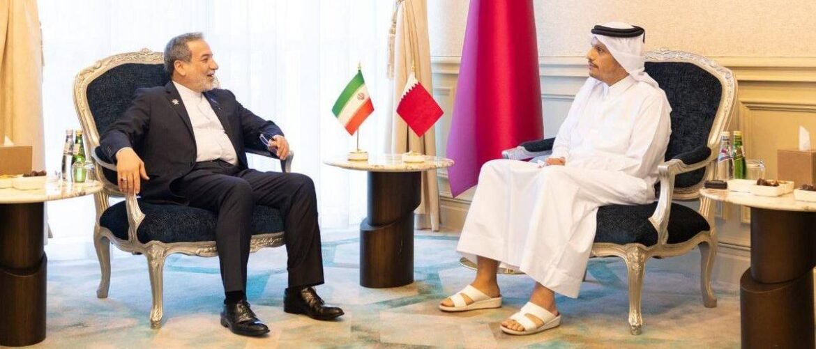 Two men in a formal meeting sit in ornate chairs across from each other, flags on a small table between them. They chat and smile in a ceremonial room.