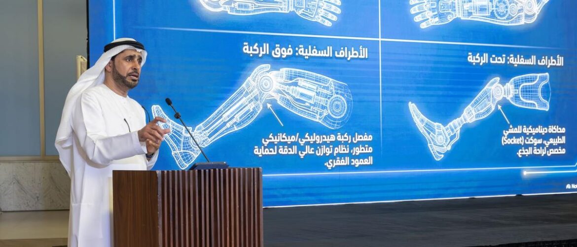Man in traditional white kandura speaks at a wooden podium during a tech presentation, with a large blue backdrop showing robotic arm diagrams in Arabic behind him.