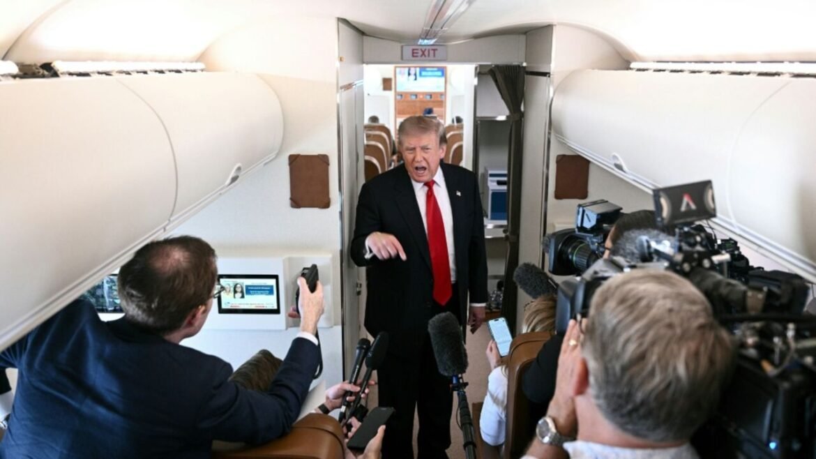 A man in a dark suit with a red tie addresses a group of reporters inside an airplane cabin, microphones aimed at him.
