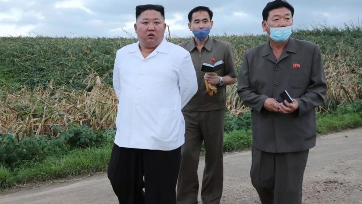 Three men stand on a dirt road beside a crop field; two wear masks and matching uniforms, one wears a white shirt.