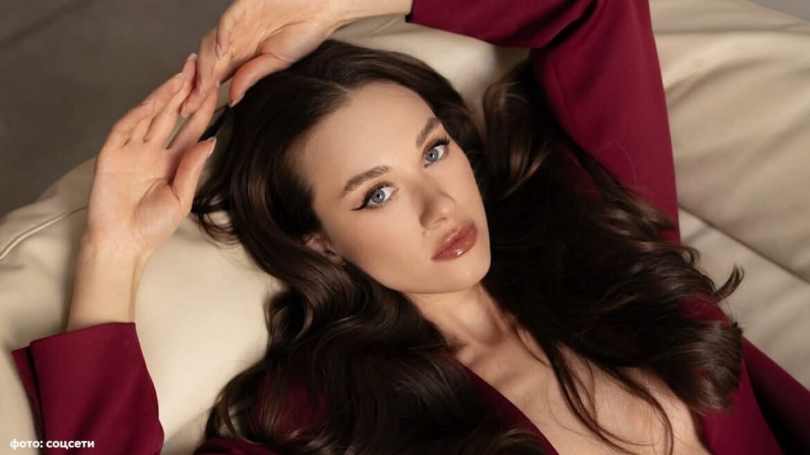 Portrait of a woman with long dark hair in a burgundy outfit, lying on a beige couch and looking at the camera.