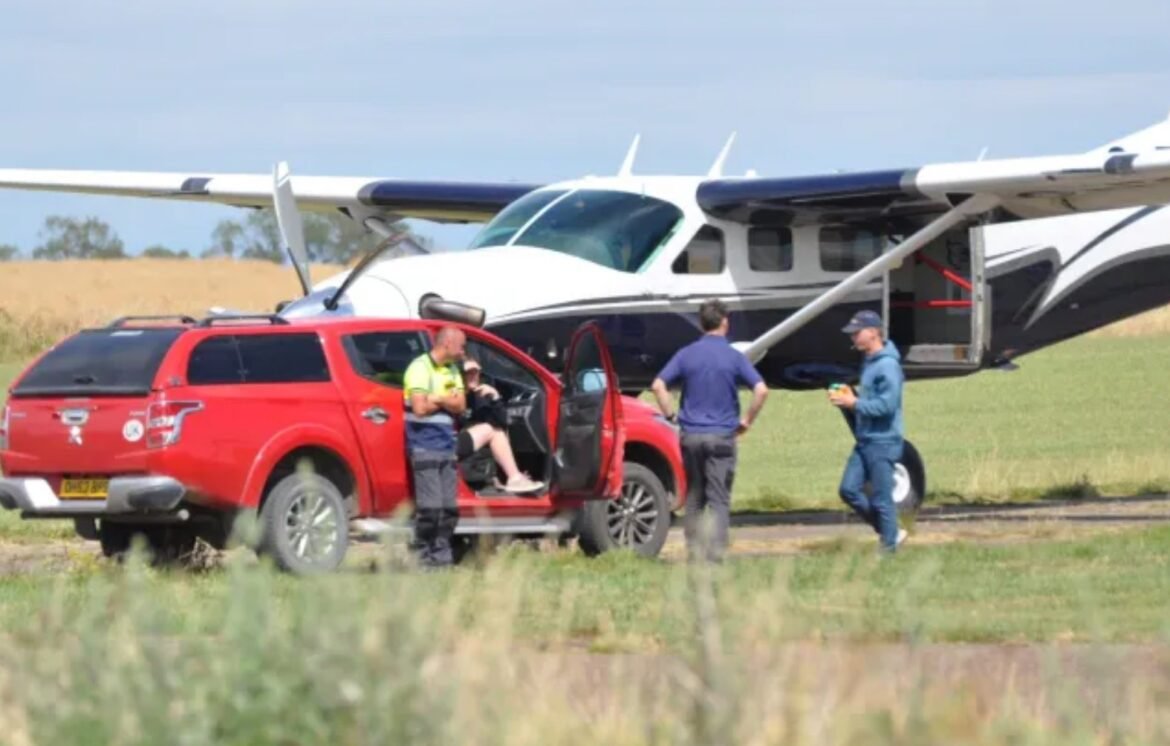 Small white and blue airplane on a grassy field with its door open; a red pickup parked beside it and three men talking nearby.