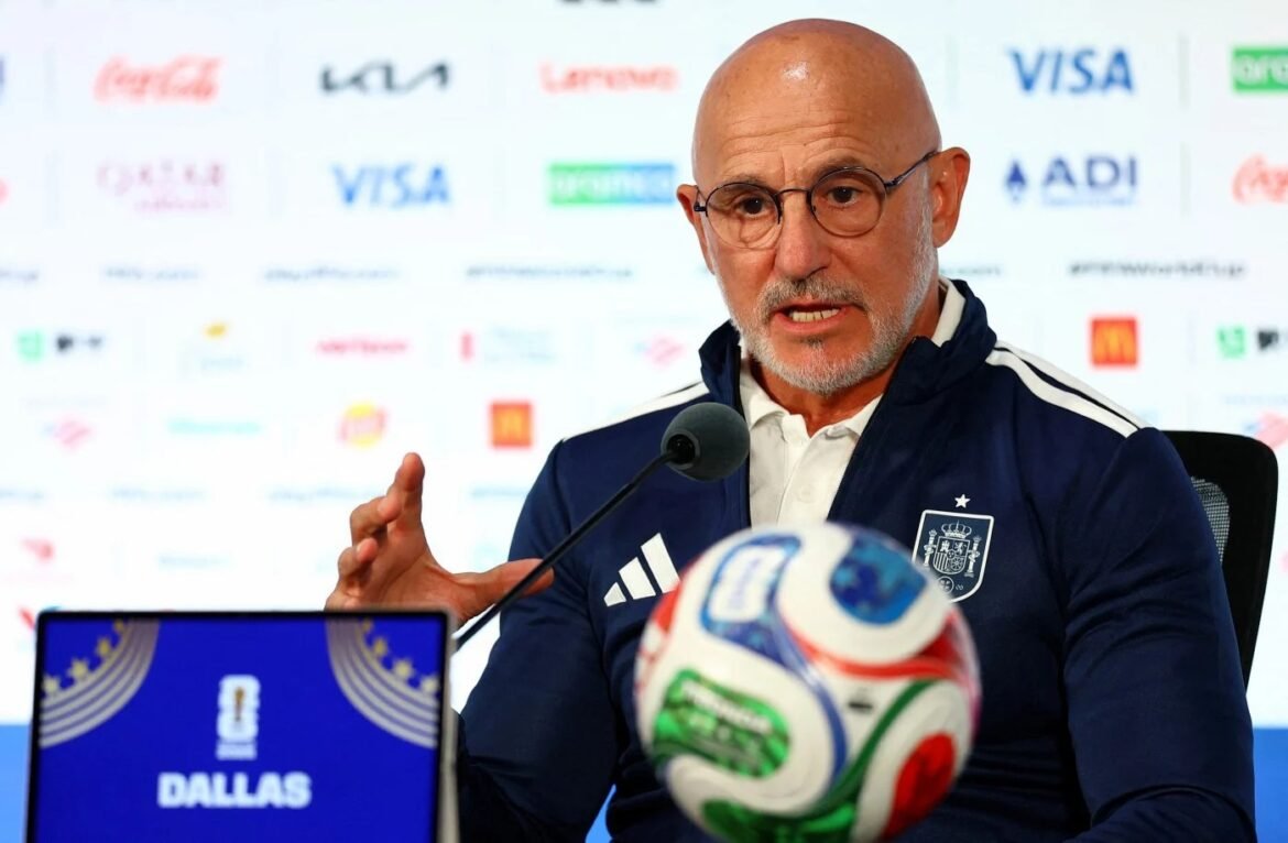 Bald man with glasses at a press conference, speaking into a microphone, wearing a blue jacket with a crest, with a colorful soccer ball nearby and sponsor backdrop behind him.
