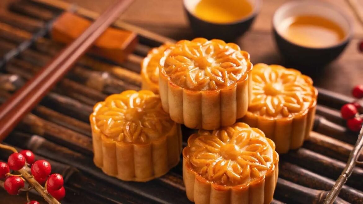 Golden-brown mooncakes with carved floral tops stacked on a metal rack, warm lighting in the background.
