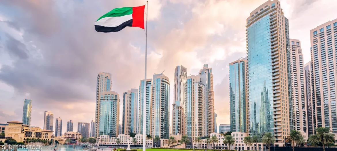 UAE flag waving on a tall pole over a modern marina skyline with glass skyscrapers.