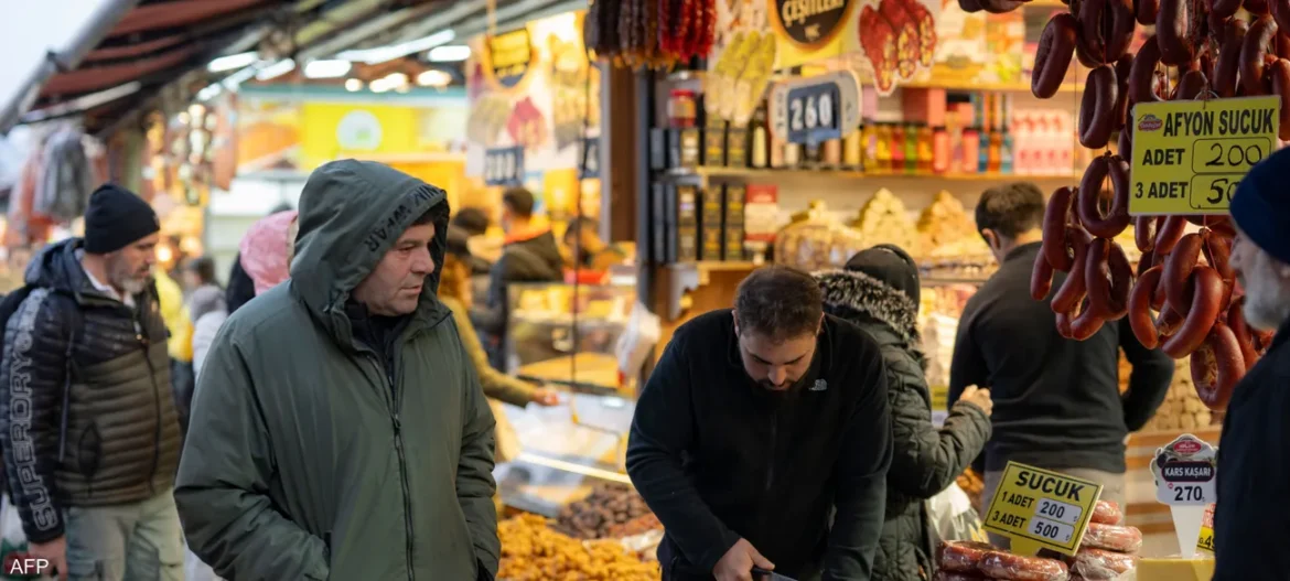 Outdoor market stall with hanging sausages on the right and shoppers browsing a deli counter in the background.