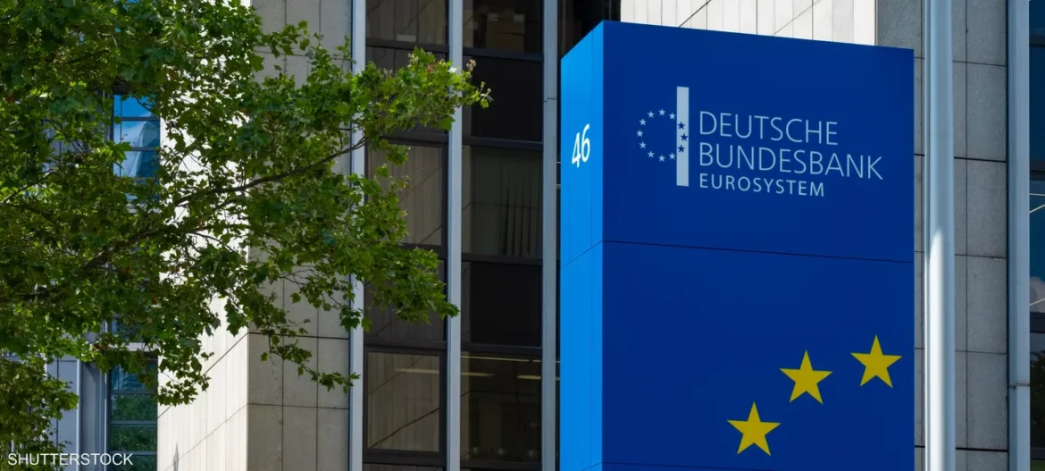 Blue sign reading Deutsche Bundesbank Eurosystem with yellow EU stars at lower right, tree on left, modern building façade in background.