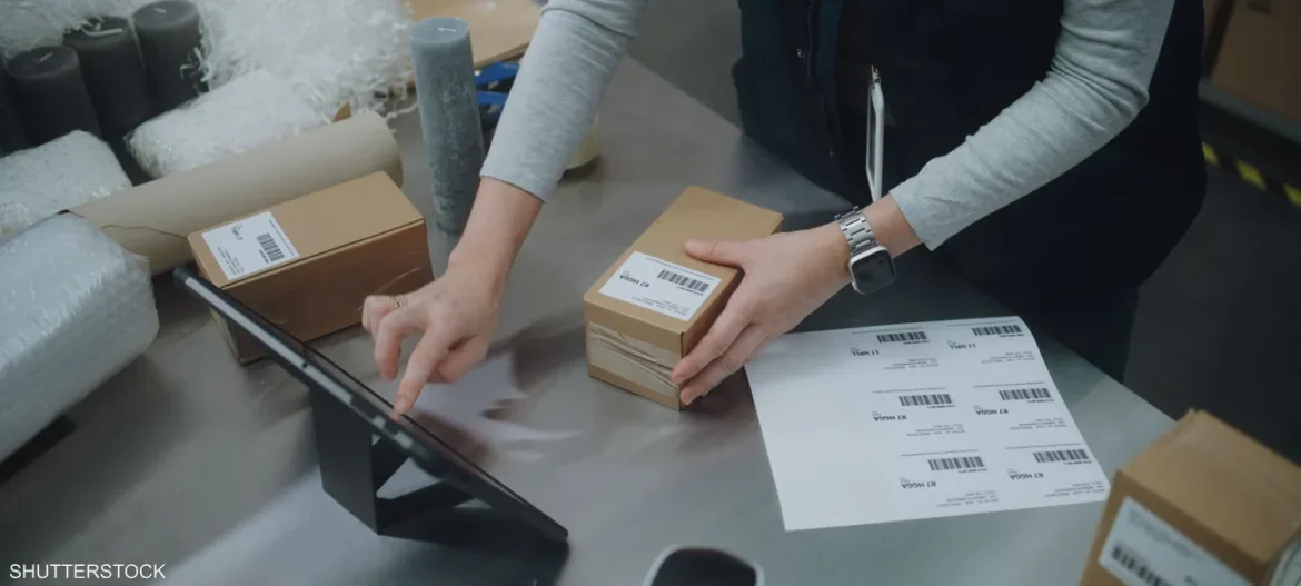 A person scans or labels small cardboard boxes at a stainless shipping desk using a tablet for order processing.