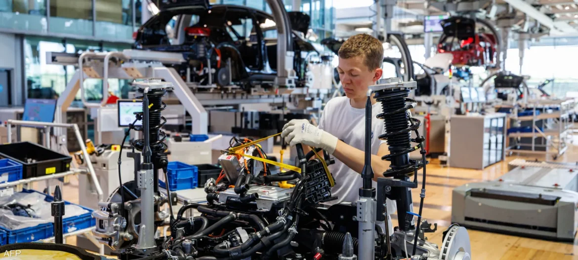 Worker in a white shirt and gloves wires an automotive chassis on a busy assembly line.