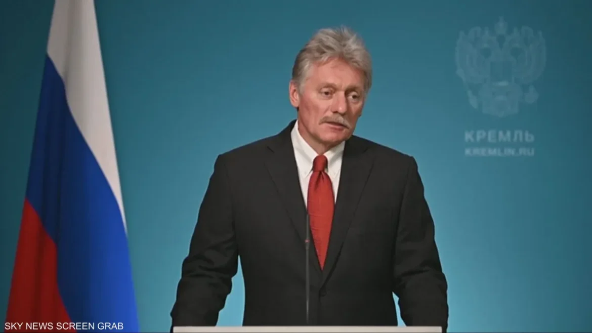 Man in a dark suit and red tie speaks at a podium, with the Russian flag visible on the left and a Kremlin backdrop behind him.