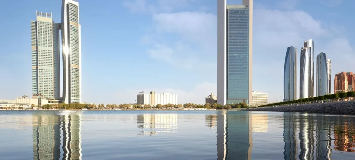 City skyline of tall glass skyscrapers along a calm waterfront with reflections in the water against a clear blue sky.