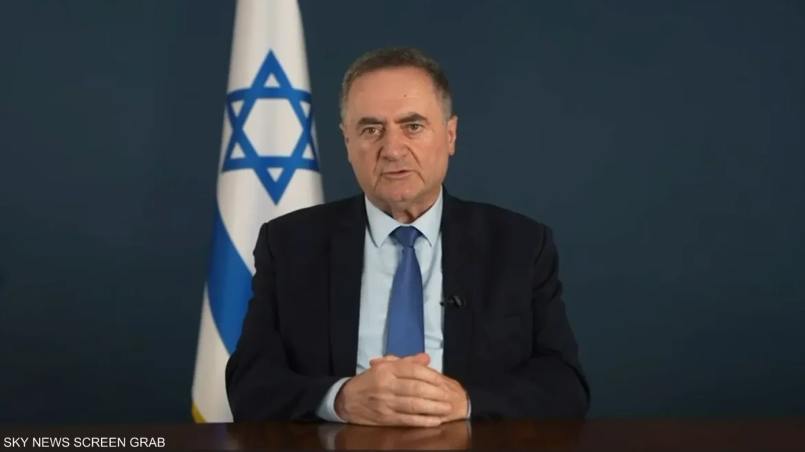 Formal video still of a man in a dark suit and blue tie speaking at a desk, with an Israeli flag visible in the background and a muted newsroom setting behind him.