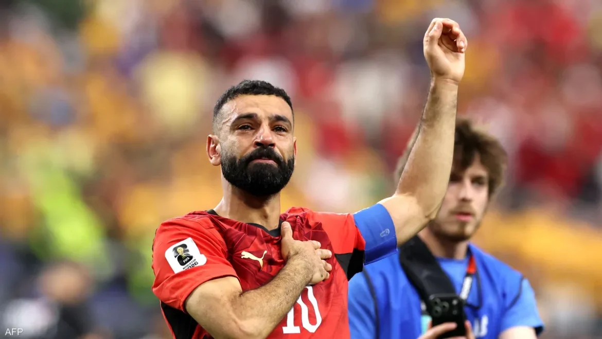 Soccer player in a red kit celebrates with a raised right arm on the field, crowd blurred in the background.