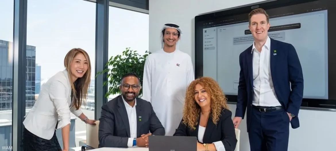 Five professionals posing for a team photo in a bright meeting room, with a laptop and large screen behind them.