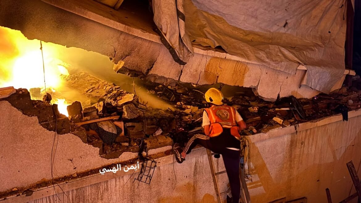 Firefighter in an orange vest and helmet climbs a ladder beside a burning, collapsed building with rubble around him.