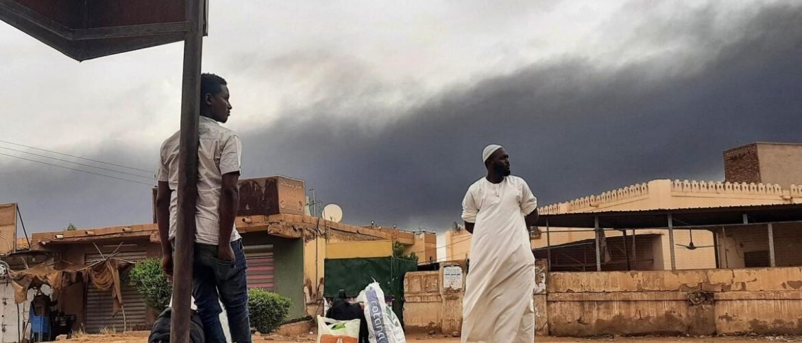 Two men stand in a dusty street under a shelter as a dark storm front approaches, one in a white robe and the other in a light shirt and jeans.